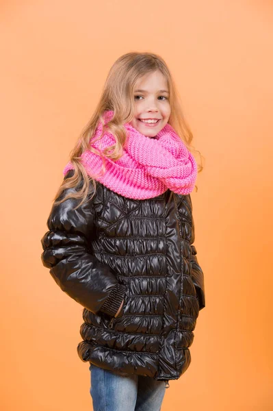 Child model smile with long blond hair. Girl in black coat and pink scarf on orange background. Autumn fashion, style, trend. Kid beauty, look, hairstyle. Happy childhood concept.