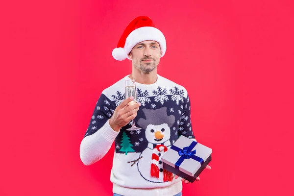 positive christmas man isolated on red background. man hold present for christmas in studio. christmas present and man with champagne. man in christmas santa hat with present and champagne.