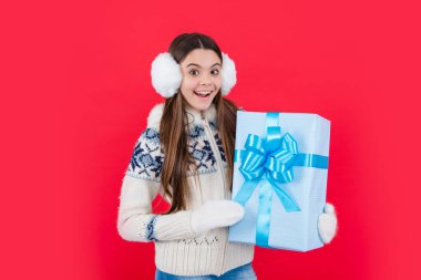 Noel tatili. Kış kulaklıklarıyla yılbaşı hediyesiyle hayrete düşmüş genç bir kız. Genç kız Noel hediyesi olarak kırmızı arka planda izole edilmiş..