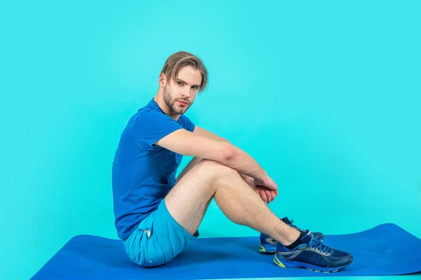 运动员坐在瑜伽垫上 与蓝色背景隔离 工作室里有一个带瑜伽垫的体育人 照片上的体育男子与瑜伽垫 穿着运动服 拿着瑜伽垫的运动员 — 图库照片