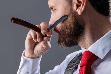 Berber bıçağıyla tıraş olan adama yakın çekim. Berber stüdyosunda berberlik yapıyor. Sakallı adam berber. Traşsız adam berber bıçak gri arka planda izole.