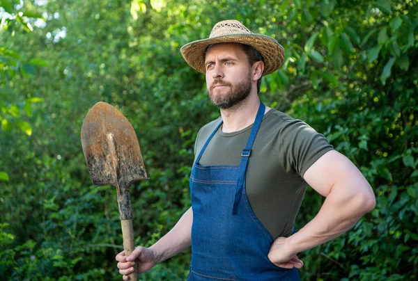 Thoughtful farmer man in farmers hat and gardening apron holding garden shovel natural outdoors.
