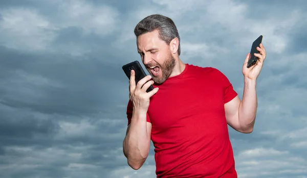 photo of angry man shouting has phone talk. man has phone talk on sky background. man has phone talk outdoor. man has phone talk wear red tshirt.