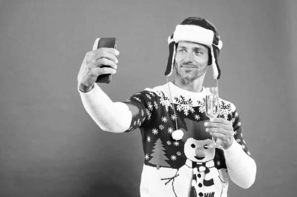 cheerful guy making selfie on smartphone on red background. express positive emotions. drinking champagne. cheers. merry christmas. celebrate new year holiday. man in winter sweater and red hat.