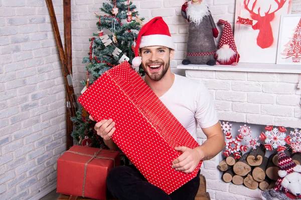 positive man with red xmas present in studio. santa man at xmas holiday. merry xmas. young man in santa hat with xmas present box.
