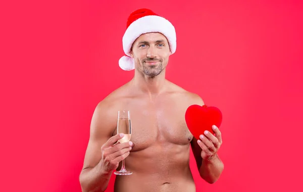 new year heart and glad man with torso. shirtless man in new year santa hat with heart and champagne. new year man isolated on red background. man hold heart at new year in studio.