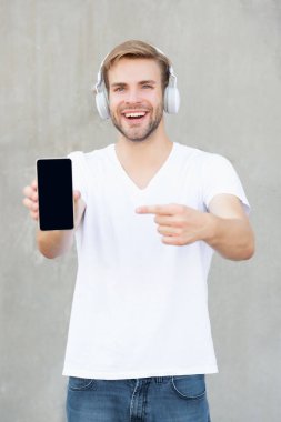 Gülümseyen adam akıllı telefondan müzik uygulamasını sunuyor. Kulaklıkla müzik uygulaması sunan bir adamın fotoğrafı. Adam müzik uygulaması sunuyor, kopyalama alanı. Adam dışarıda müzik uygulaması sunuyor..