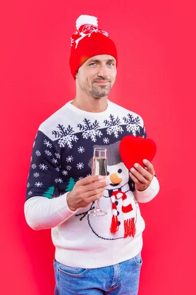 man in new year hat with champagne and heart. new year man isolated on red background. man hold champagne for new year eve in studio. new year heart gift and man in sweater.