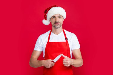 Kırmızı Noel Baba şapkalı ve önlüklü olgun bir adam kırmızı arka planda mutfak bıçağı taşıyor..