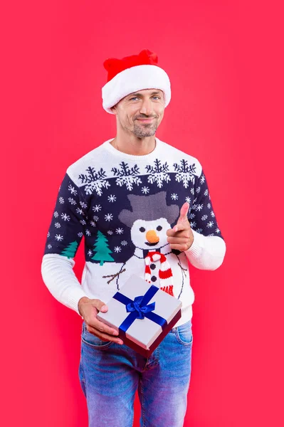 man hold present for xmas pointing finger in studio. xmas present and man in sweater. man in xmas santa hat with present box. xmas man isolated on red background.