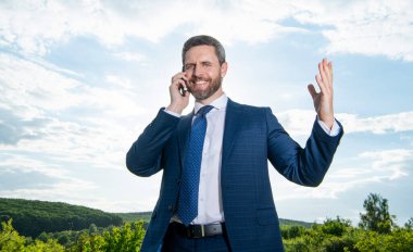 Gülümseyen adam dışarıda telefonla konuşuyor. Adamın telefonda konuşma kıyafeti var. Adamın fotoğrafı telefonda konuşuluyor. Adamın gökyüzü geçmişinde telefon konuşması var..