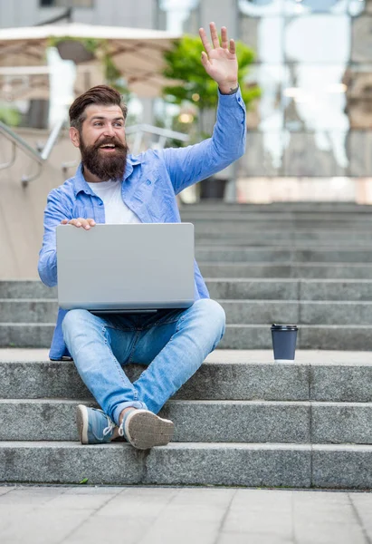 Happy freelancer making hey gesture sitting on stairs. Male freelancer working online outdoors. Freelancer freelancing online. Freelancer man using laptop computer for remote work online.