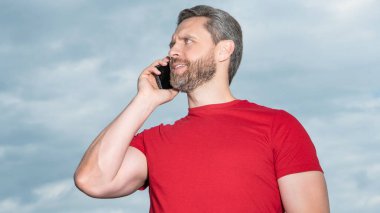 Mutlu adamın gökyüzü arka planında telefon konuşması var. Adam dışarıda telefonla konuşuyor. Adam telefonda konuşuyor, kırmızı tişört giyiyor. Bir adamın fotoğrafı telefonda konuşuluyor..