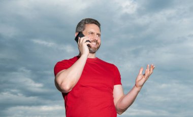 smiling man has phone talk on sky background. man has phone talk outdoor. man has phone talk wear red tshirt. photo of man has phone talk.