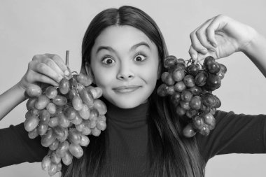 Şaşırmış pozitif genç kız sarı arka planda bir demet üzüm tutuyor.