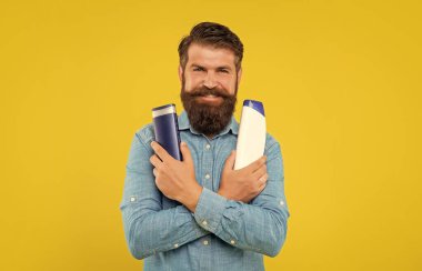 Gülümseyen vahşi sakallı adam sarı arka planda saç kremi şişesini seçiyor ve tuvalet malzemelerini sunuyor..