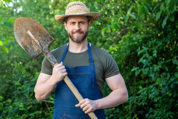 Happy gardener man in gardening apron and farmers hat holding garden spade natural outdoors.