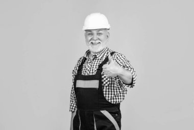 Sarı arka planda kasklı, yaşlı ve mutlu bir inşaatçı. başparmak yukarı.