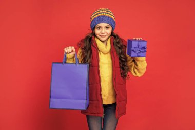 Örgü şapkalı neşeli genç kız alışveriş çantası ve kırmızı arka planda hediye kutusu, alışveriş.