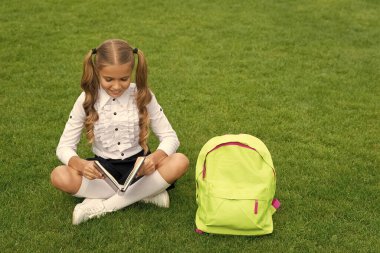 Okul çantalı mutlu kız çocuğu yeşil çimlerin üzerinde oturmuş kitap okuyor..
