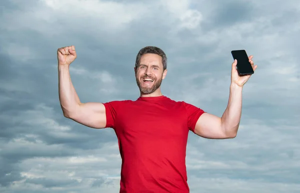 successful man showing phone app wear red tshirt. photo of man showing phone app. man showing phone app on sky background. man showing phone app outdoor.