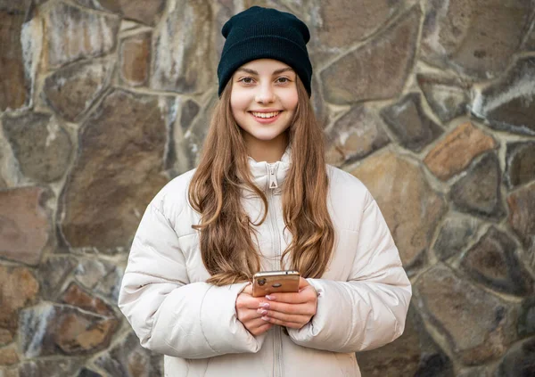 photo of smiling teen girl blogging on phone outside. teen girl blogging on phone at stone background. teen girl blogging on phone outdoor. teen girl blogging on phone wearing hat.