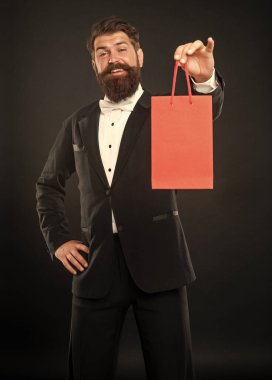 Smokinli mutlu bir adam siyah arka planda papyon takıyor ve erkekler günü için alışveriş çantası sunuyor. Boşluğu kopyala.