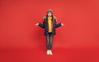 Kış şapkası ve kırmızı arka planda ceketi olan kafası karışmış bir çocuk..