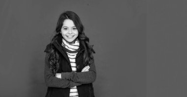 Kabarık yelekli kıvırcık saçlı neşeli çocuk. Kırmızı arka plandaki genç kız. Kalın giysiler giyen çocuk portresi. Olumlu duygularını ifade et. Kış modası. Boşluğu kopyala.