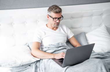 Yatakta dizüstü bilgisayarda çalışan gözlüklü yetişkin işveren..