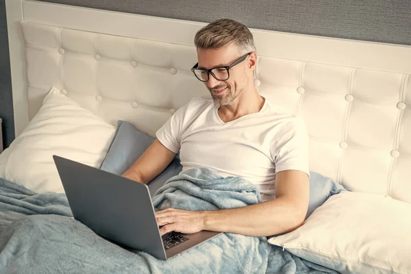 Yatakta dizüstü bilgisayarda çalışan gözlüklü olgun bir adam..