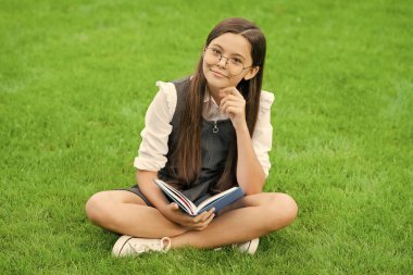 Gözlüklü neşeli çocuk yeşil çimenlerde oturmuş kitap okuyor..