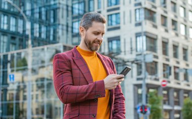 cheerful man messaging on smartphone in the street. man messaging on smartphone outside. photo of man messaging on smartphone in jacket. man messaging on smartphone outdoor.