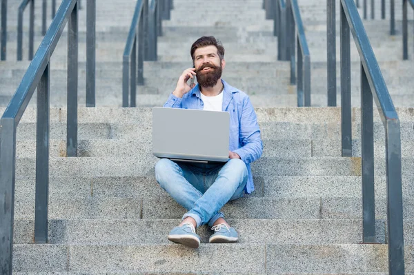 glad guy has online video communication sitting on stairs. guy has online video communication on laptop. guy has online video communication outdoor. guy has online video communication outside.