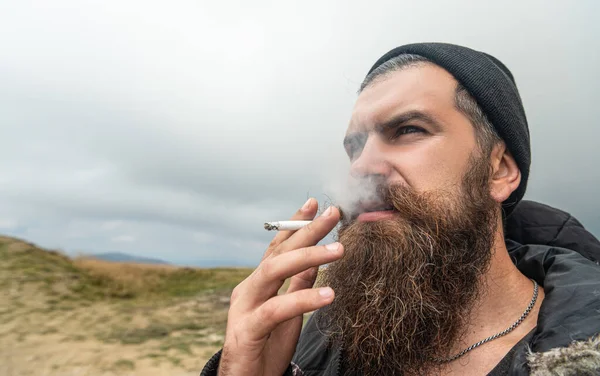caucasian bearded man smoking cigarette. photo of man smoking cigarette in the mountain. man smoking cigarette outdoor. man with beard smoking cigarette.