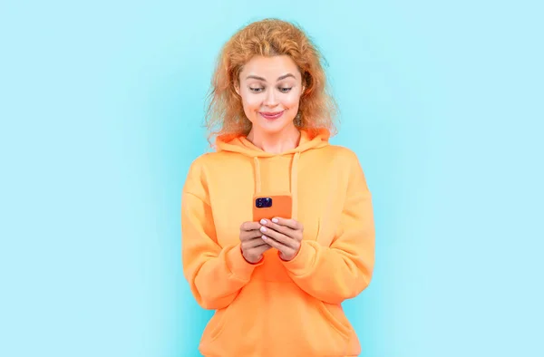 photo of cheerful woman chat on smartphone in hoodie. redhead woman chat on smartphone on color background. woman chat on smartphone isolated on blue background. woman chat on smartphone at studio.