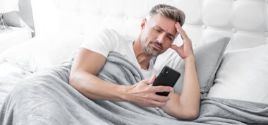 Uykulu, olgun bir adam yatakta telefonda konuşuyor..