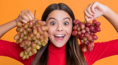 Şaşırmış genç kız sarı arka planda bir demet üzüm tutuyor.