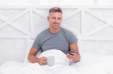 Mutlu bir adam sabah çayıyla yatakta dinlenirken akıllı telefondan mesaj atıyor, mesajlaşıyor..