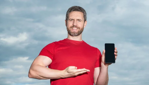 man presenting phone app outdoor. man showing phone app wear red tshirt. photo of man showing phone app. man showing phone app on sky background.