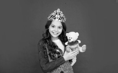 Kraliçe tacındaki gülümseyen çocuk. Tacın içindeki prenses. Çocuk ayı oyuncağını tutuyor. Genç kız kırmızı arka planda diyadem giyiyor. Mutlu çocukluk.