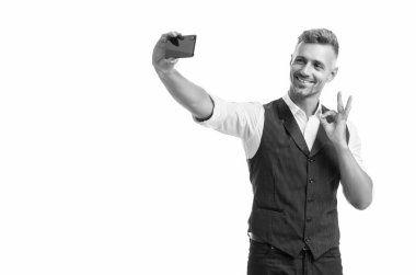Takım elbiseli mutlu yönetici, beyaz renkte izole edilmiş akıllı telefonlu selfie çekerken yüzük jesti gösteriyor..