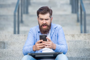 Upset man checking up mobile phone betting results while sitting on steps outdoors. Betting using mobile apps in smartphone. Betting online on mobile device. Mobile betting. Making bet, gambling.