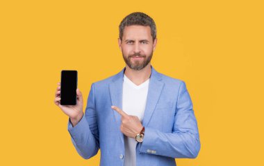 businessman pointing on portable phone isolated on yellow. businessman hold portable phone in studio. businessman showing portable phone. businessman presenting screen of portable phone