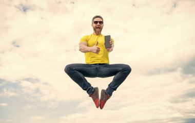 man jumping and presenting smartphone on sky background. thumb up.