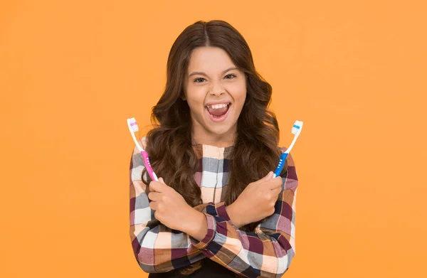 Teeth whitening. Girl cute long hair holds toothbrushes yellow background. Child girl holds two toothbrushes. Child school girl smart kid happy face cares hygiene. Brush teeth concept. Teeth hygiene.