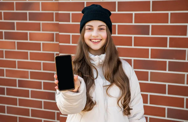 girl presenting phone app outdoor, copy space. girl presenting phone app wearing hat. photo of girl presenting phone app outside. girl presenting phone app on brick background.