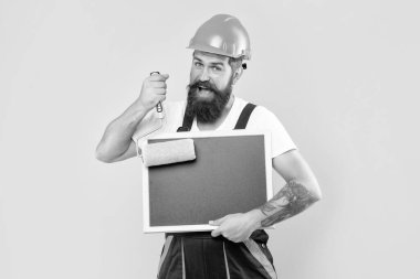 Kasklı ve iş elbiseli gülümseyen adam sarı arka planda fotokopi alanı olan boya silindiri ve karatahta tutuyor..