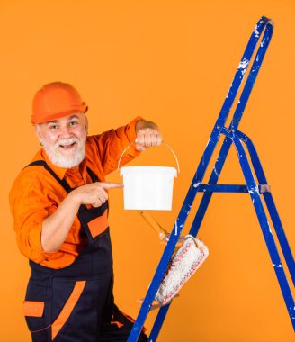 Boyalı kovayla şapkalı bir ressam. İnşaat işçisi boyama cephesi. Isı yalıtımı işe yarıyor. Boyacı merdiveni. Onarım yapmak için. İş elbiseli bir adam boş odada duvar boyuyor..