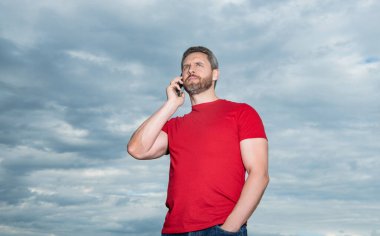 bearded man has phone talk wear red tshirt. photo of man has phone talk. man has phone talk on sky background. man has phone talk outdoor.